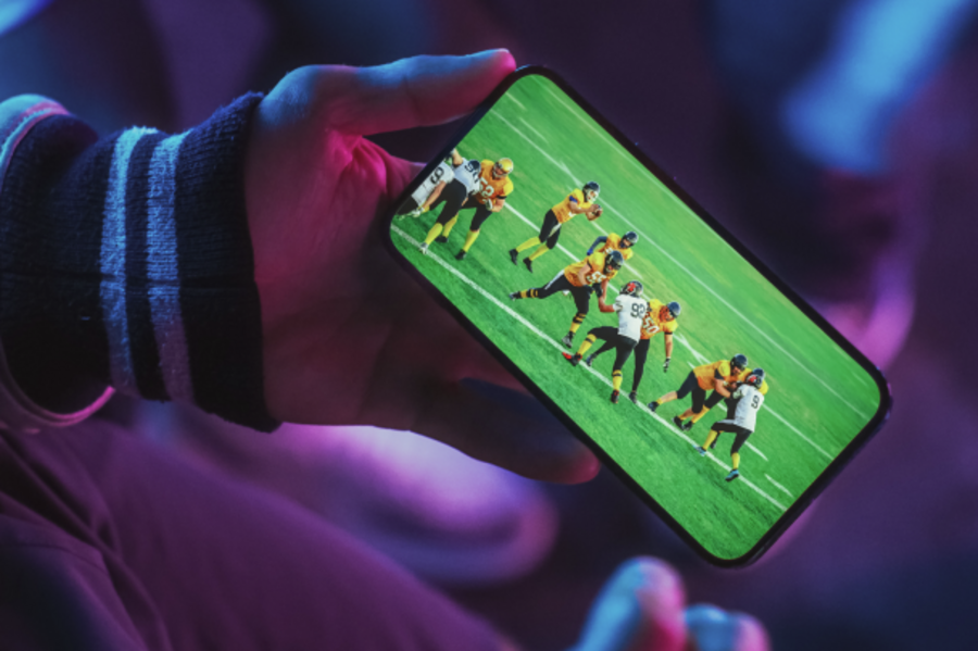 Eine Person hält ein Smartphone in der Hand und schaut darauf eine Live-Sportübertragung eines American-Football-Spiels.