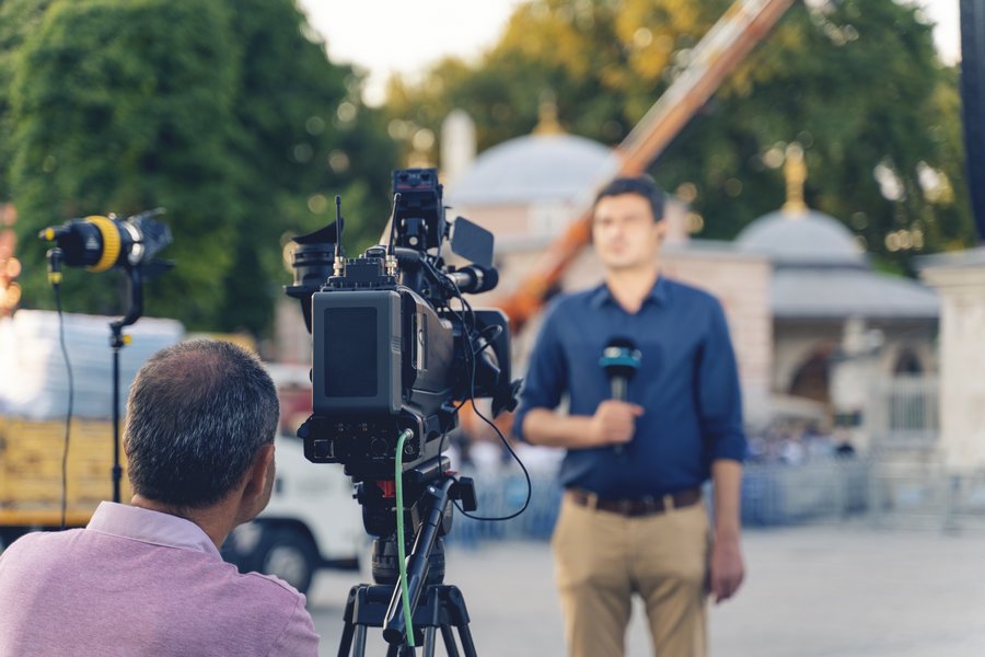 Camera crew filming a reporter during a live broadcast outdoors in front of a historic building.